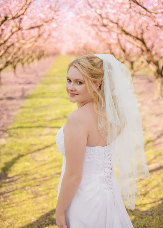 Bride looking back in a blooming orchard aisle, color correction and soft color grading, skin retouching with natural texture