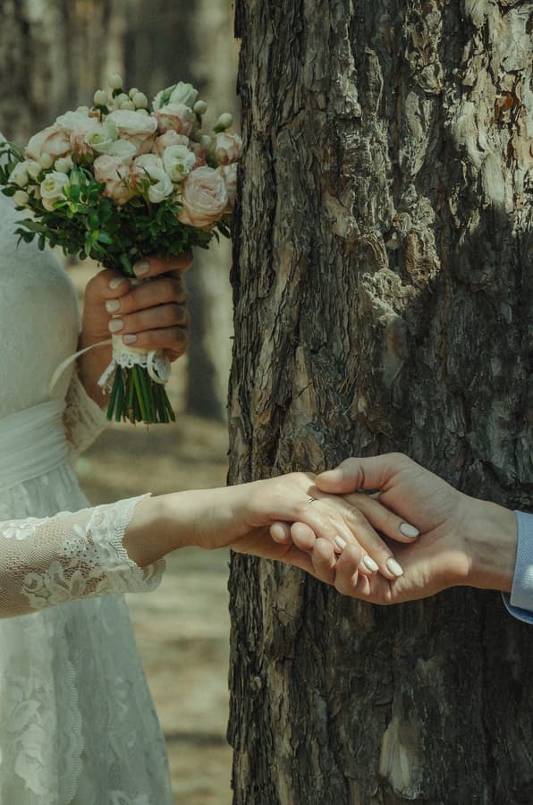 Bride and groom holding hands near a tree with wedding bouquet, skin retouching with natural texture, color correction for warm tones