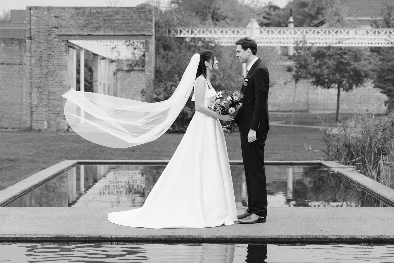Black and white photo of bride and groom standing by a reflecting pool with veil flowing in the wind